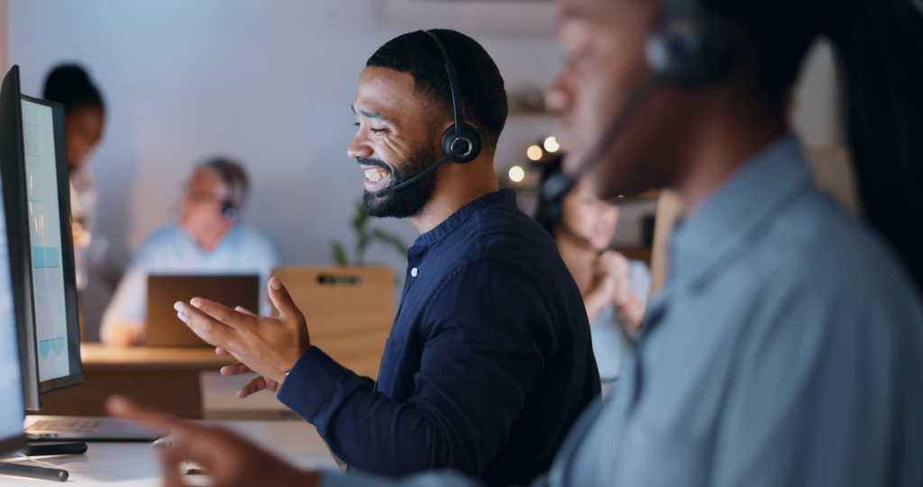 A person wearing a headset, smiling and talking while looking at a computer screen, appears engaged in help desk support services. Another individual is blurred in the foreground, while two more colleagues diligently work in the background of the bustling office setting.