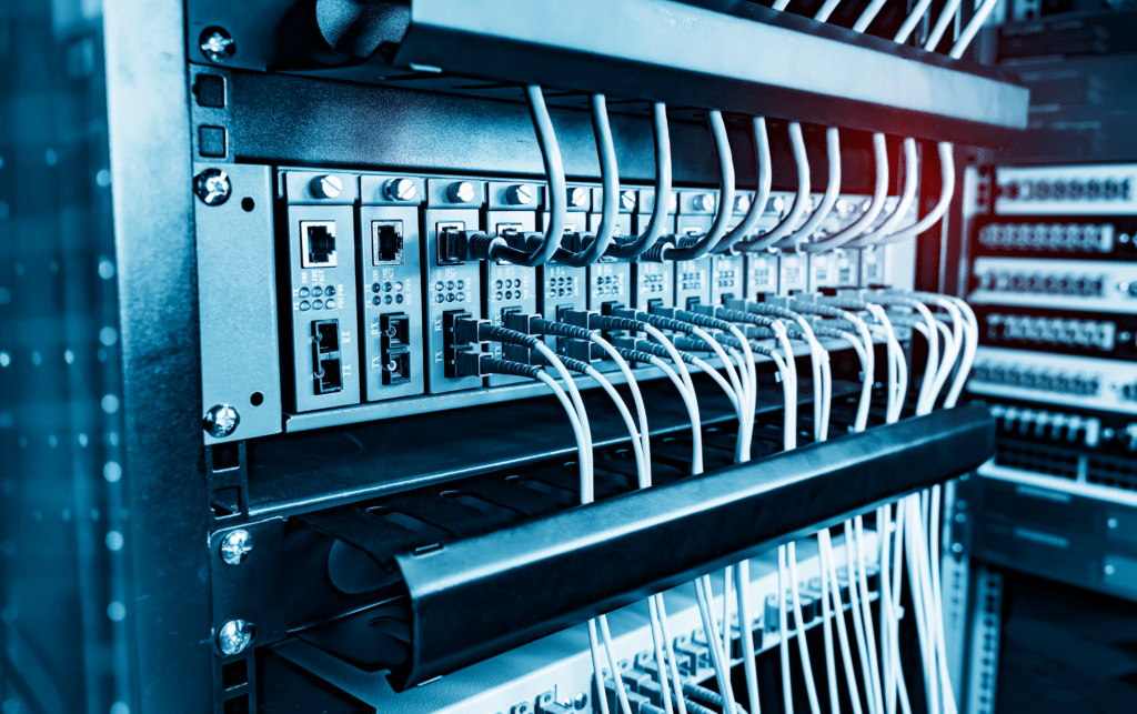 A close-up of a server rack with multiple network cables connected to various ports, showcasing managed network solutions. The cables are neatly organized, and the equipment is arranged in a dark metallic frame, glowing slightly from ambient blue lighting.