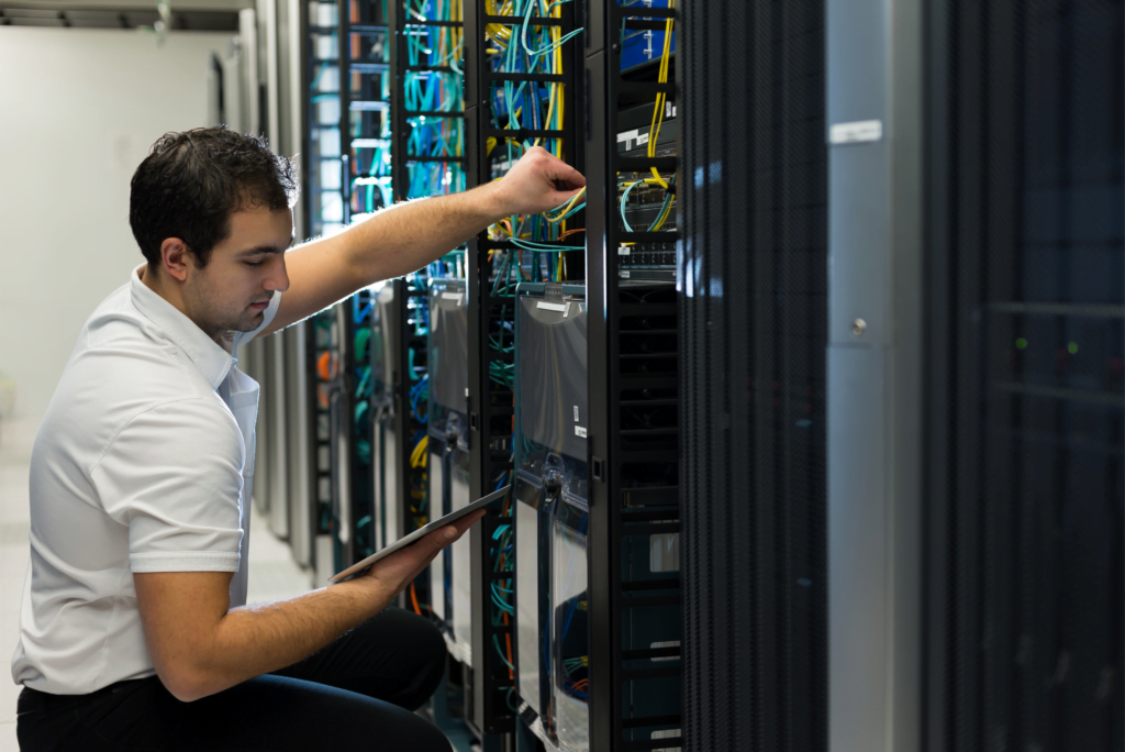 In a bustling data center, a man in a white shirt kneels before a server rack, showcasing the efficiency of managed network solutions. With a tablet in one hand and adjusting cables in the other, he navigates through rows of intricate network equipment and cables.