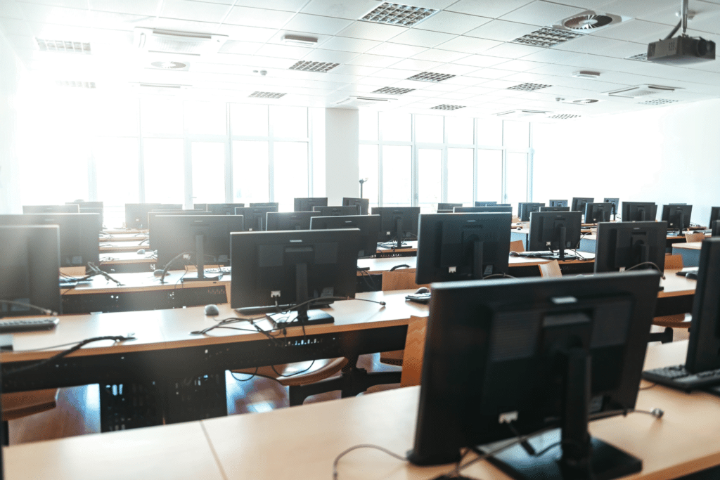 A bright computer lab with multiple rows of wooden desks and chairs, each equipped with a desktop computer and monitor. Large windows line the far wall, allowing natural light to illuminate the room, creating an efficient space for IT asset management solutions.