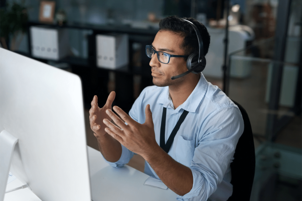 A man wearing glasses and a headset is sitting at a desk, gesturing with his hands in front of a large monitor. He appears to be discussing IT Asset Management solutions during a video call or virtual meeting in a modern office setting.