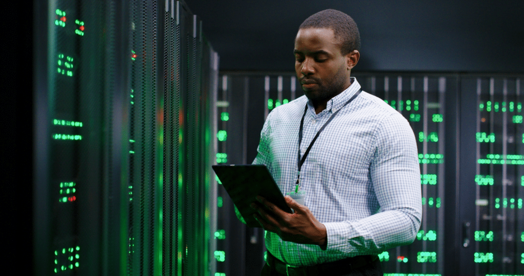 A person in a checkered shirt stands in a dimly lit server room, holding a tablet. The servers, critical to managed disaster recovery, emit green lights as they diligently work. The atmosphere is focused and high-tech.