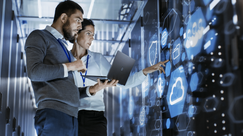 Two people stand in a server room, examining a virtual display with cloud computing icons. One holds a laptop while the other points at the holographic interface showcasing cloud migration solutions and data symbols. They are focused and engaged.