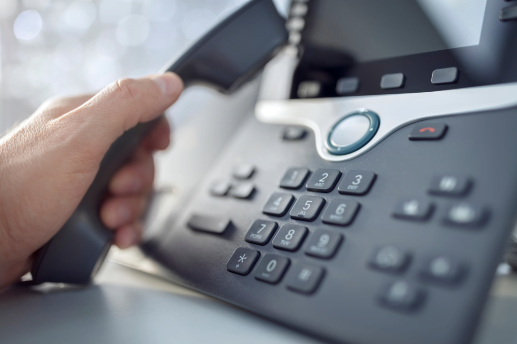 A close-up of a hand picking up the receiver of a modern desk phone, seamlessly integrating Managed VoIP solutions. The device features a digital display and keypad with various buttons, while the blurred background emphasizes the phone and hand as the primary focus.