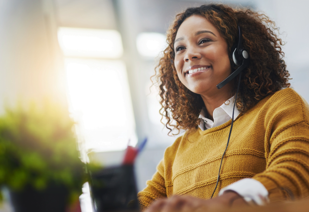 A smiling woman in a yellow sweater, representing managed VoIP solutions, sits at a desk with her headset on, engaging with her computer screen. A plant decorates the bright and sunny setting.