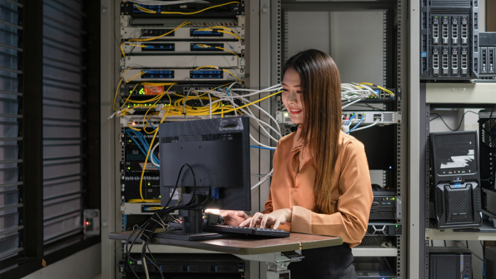 A person in a brown blouse stands in a server room, using a computer. Surrounded by equipment racks and wires, they seamlessly integrate managed server solutions into the tech-focused environment.