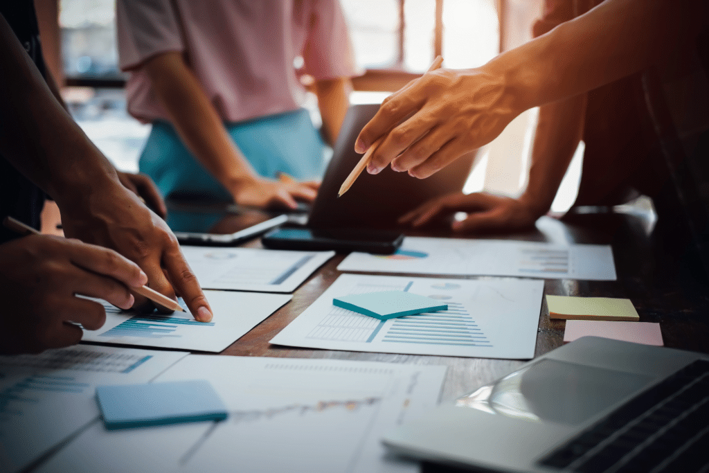 A group of people collaborating at a table covered with papers, graphs, and Post-it notes. They're using pencils to point at charts and discuss VCIO solutions, with laptops nearby, suggesting a business meeting or brainstorming session focused on innovative VCIO services.