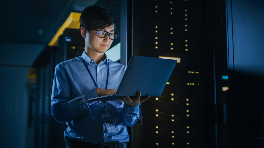 In a dimly lit server room, a person wearing glasses and a blue shirt is diligently working on a laptop, surrounded by glowing server racks, ensuring data backup and disaster recovery processes are flawlessly executed.