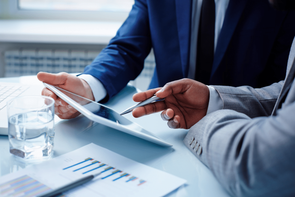 Two individuals in business attire are engaged in a discussion over a tablet at a desk, one holding a pen. A glass of water and documents with graphs are visible, emphasizing the professional cloud consulting setting.