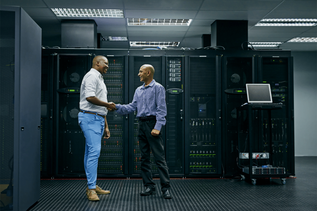 In a brightly lit server room, two men exchange a handshake, surrounded by tall black server racks. One dons a white shirt and blue pants, the other in blue and dark pants. Nearby, a laptop on a stand signifies the presence of their 24/7 managed SOC operations.