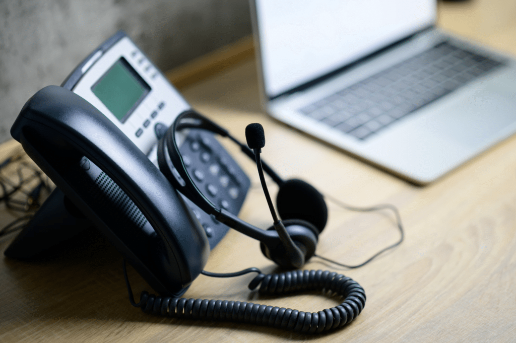 A desk features a laptop with its screen slightly open, accompanied by a business phone and headset. The digital display phone, essential for VoIP services for business, has buttons ready for action. The headset, with an attached microphone, rests casually on the handset.