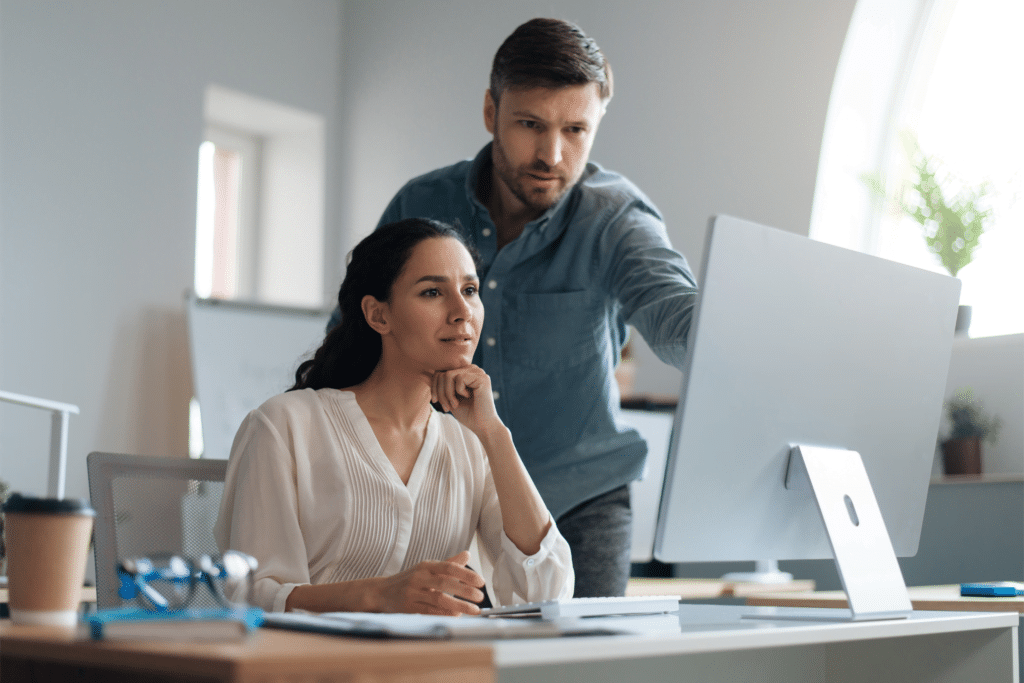 A man and woman are in an office, focused on a computer screen. The woman, pen in hand, is seated thoughtfully as the man points out details about endpoint security solutions. Nearby lie glasses and a notepad on the desk, adding to the air of diligent problem solving.