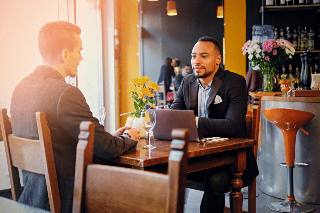 Two men in business attire sit across from each other at a restaurant table with a laptop open, discussing IT managed services. One man has a notepad. The setting is cozy, with warm lighting, flowers, and a bar in the background.