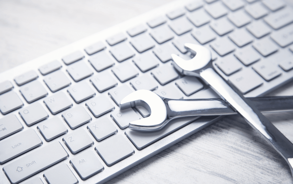 A white computer keyboard with two metal wrenches placed diagonally on top illustrates the concept of MSP vs MSSP in technical repair or computer maintenance. The setting rests on a light-colored, wooden surface, enhancing the juxtaposition of technology and tools.