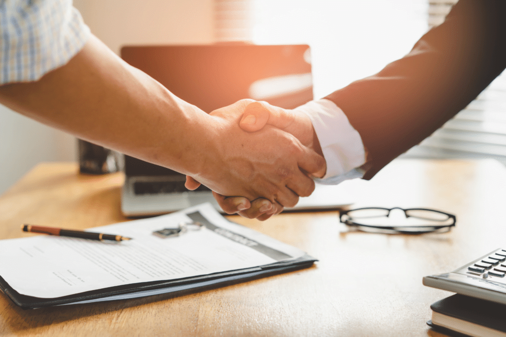 Two people are shaking hands over a wooden desk, perhaps finalizing a deal about MSP vs MSSP services. A laptop, pair of glasses, pen, and document lie neatly on the desk. Sunlight filters through the blinds in the background, casting a warm glow over this professional moment.