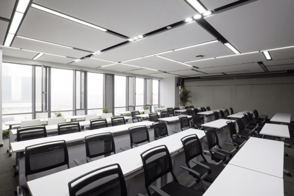 A modern, well-lit conference room with rows of long white tables and black mesh office chairs. Large windows on one side provide natural light, complemented by green plants along the windowsills. The space is ideal for discussions on addressing the cybersecurity skills gap. Geometric lighting graces the ceiling.