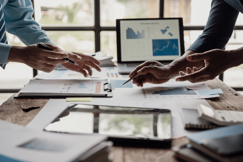 Two people discussing business charts and graphs at a wooden table with a laptop, perfecting their IT setup for startups. Hands gesturing over documents highlight collaboration and data analysis in this modern office setting.