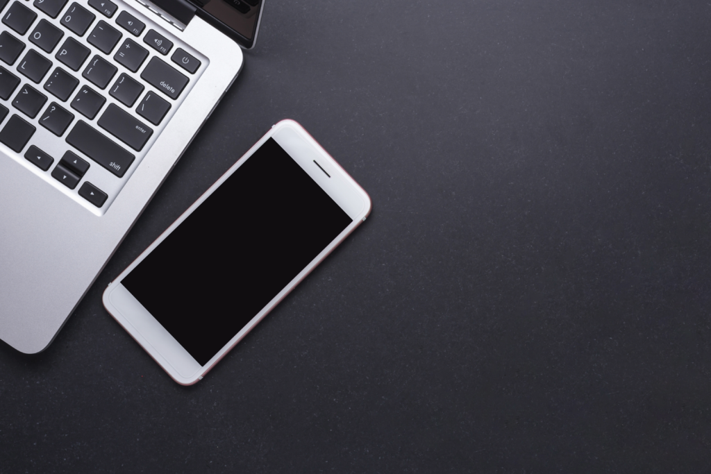 A silver laptop with part of its keyboard visible sits on a dark surface next to a white smartphone, hinting at Mobile Device Management: Enhancing Security and Productivity. The devices are in the upper left, leaving space on the right.