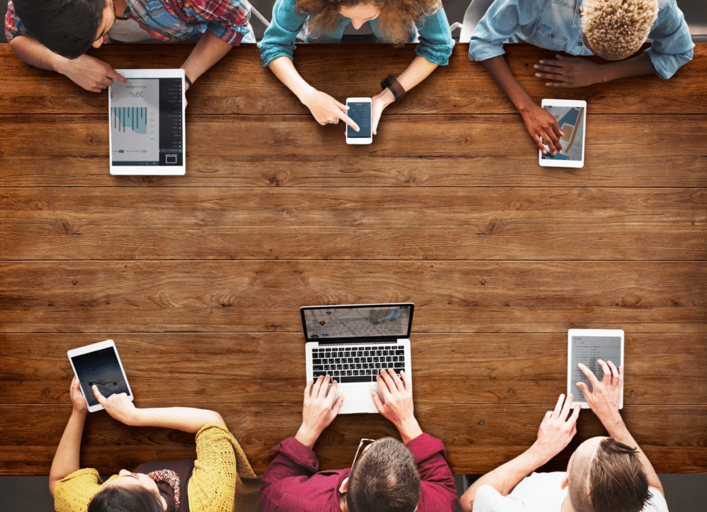 A diverse group of people sits around a wooden table, using various electronic devices like laptops, tablets, and smartphones in a collaborative meeting. Mobile Device Management plays a key role here, enhancing security and productivity as they engage in their tasks.