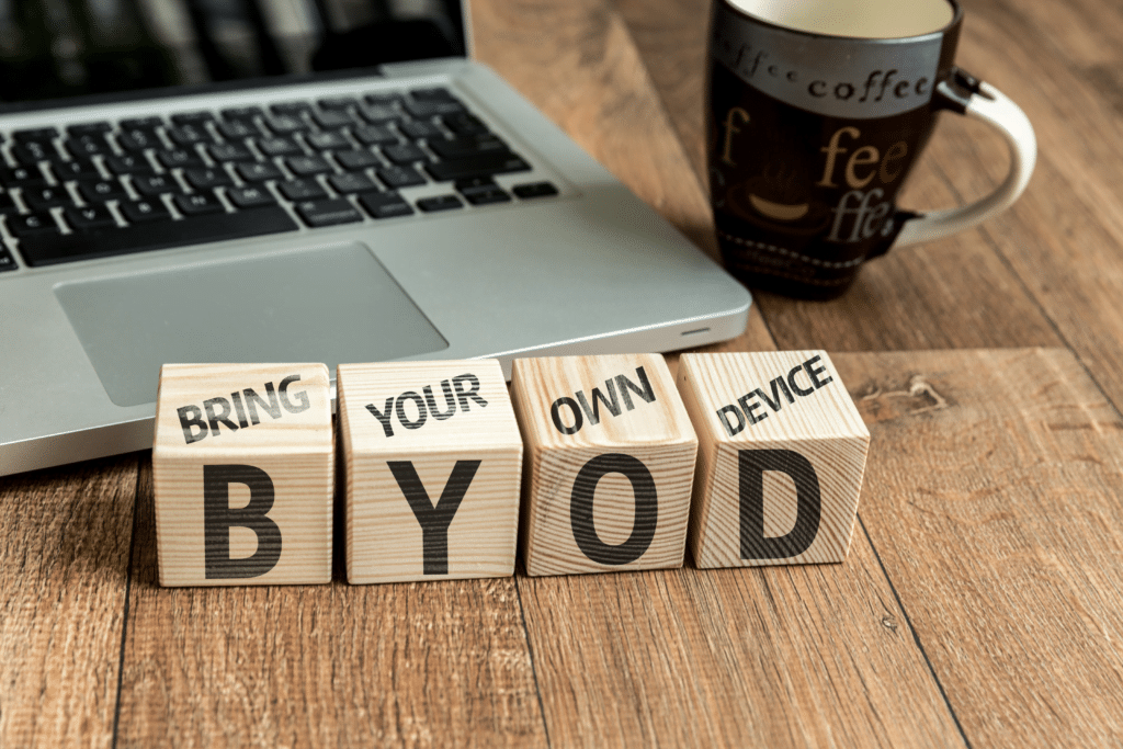 Wooden blocks on a desk spell out BYOD with Bring Your Own Device written underneath, highlighting the importance of Mobile Device Management in enhancing security and productivity. A laptop and a coffee mug are visible in the background on a wooden surface.