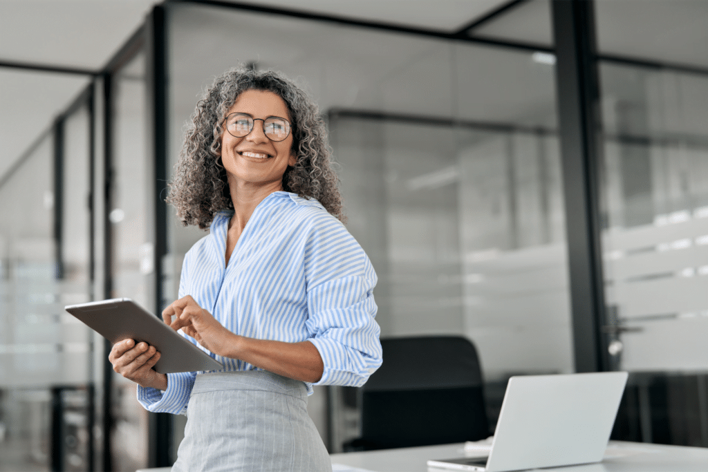 A smiling woman with curly hair and glasses stands in an office, holding a tablet. She wears a striped shirt and gray pants. A laptop rests on the desk behind her, hinting at the dynamic industries we serve, with glass walls visible in the background.