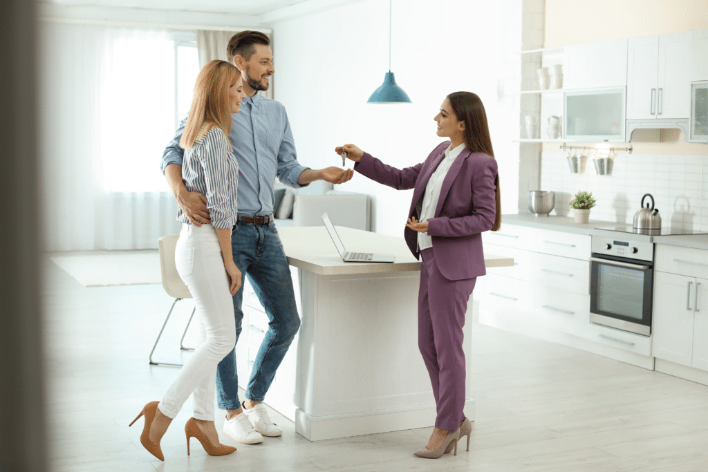 In a bright, modern kitchen, a couple stands with a real estate agent in a purple suit who hands them keys. The agent smiles warmly, nodding towards the laptop on the island, highlighting the diverse industries they serve in their journey to find dream homes.