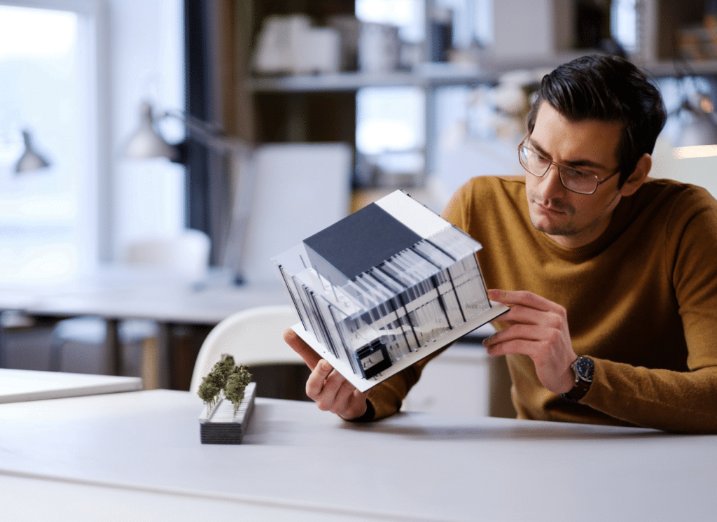 A man wearing glasses and a brown sweater examines a modern architectural model in a bright office, highlighting the industries we serve. A small model of trees is on the table in front of him, with shelves and workspaces in the background.