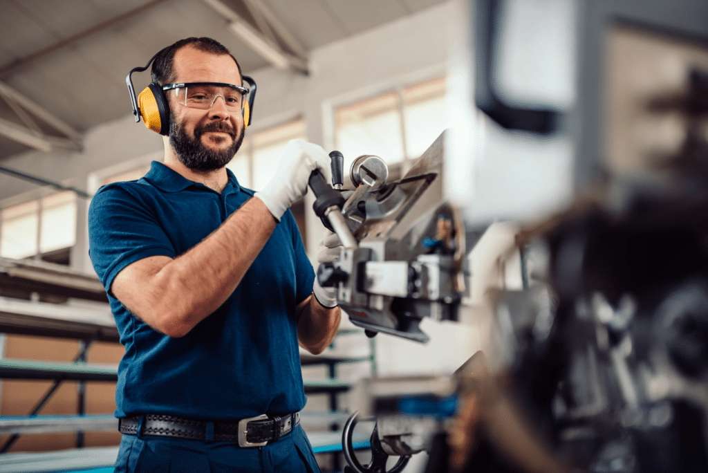 A man wearing safety glasses, ear protection, and gloves operates a metalworking machine in an industrial workshop. He smiles slightly while adjusting the machine, surrounded by various tools and equipment found across the industries we serve.