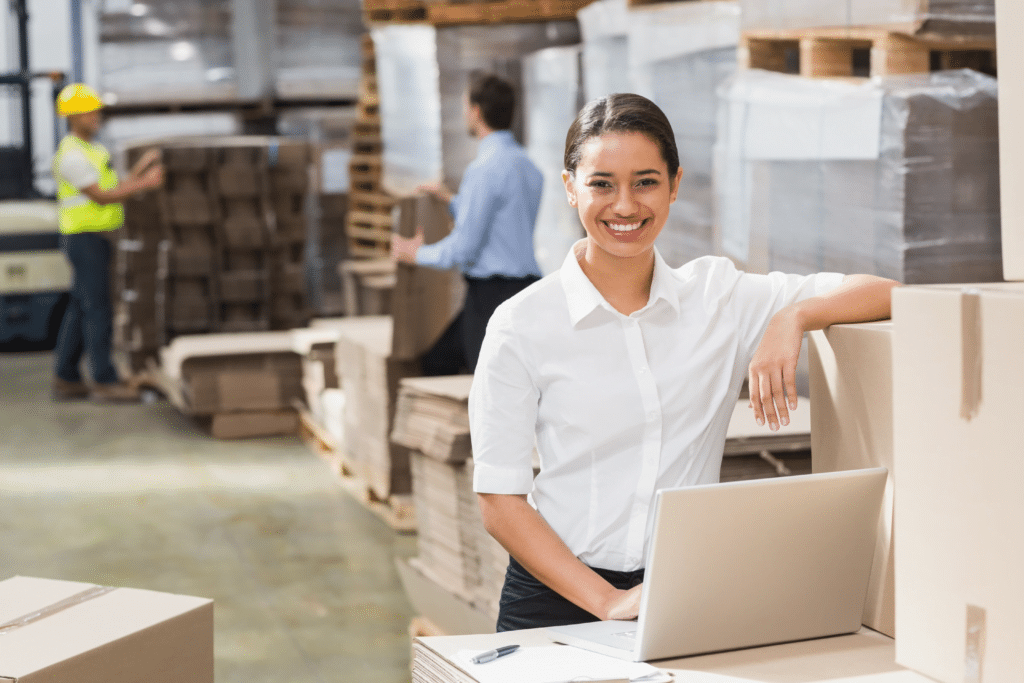 A woman in a white shirt smiles while leaning on stacked boxes and working on a laptop in a bustling warehouse, showcasing the diverse industries we serve. In the background, a worker in a yellow hard hat handles more boxes, surrounded by shelves filled with packages.