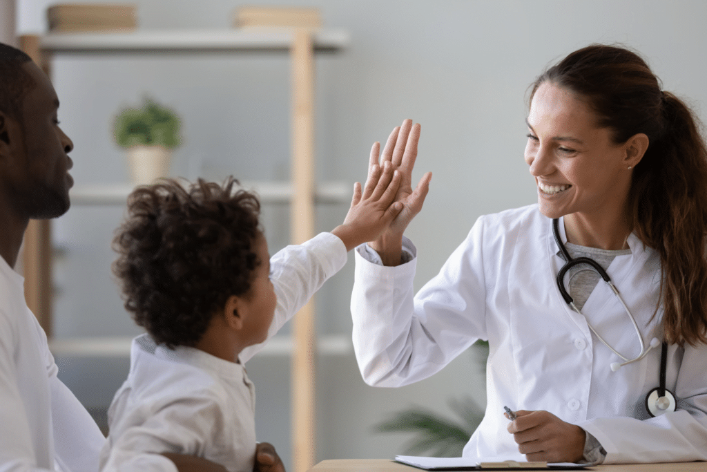 A doctor in a white coat smiles while giving a high-five to a young child seated at a table. An adult sits nearby, partially visible. The setting appears to be a medical office, showcasing one of the many industries served by healthcare professionals with shelves in the background.