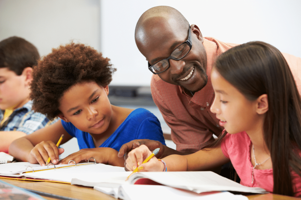 A teacher smiles while assisting two students with their work in a classroom, showcasing the dedication seen across the diverse industries we serve. The focused students hold pencils, surrounded by open notebooks and textbooks, as others engaged in learning are visible in the background.