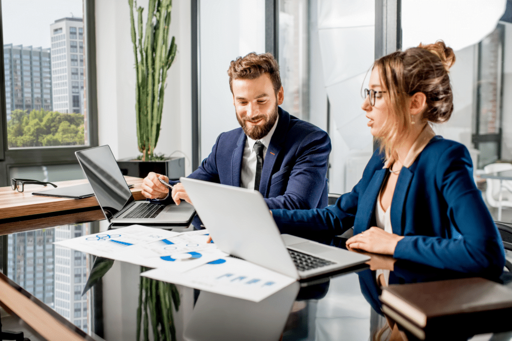 A man and woman in business attire work on laptops at a conference table, surrounded by papers and charts detailing the various industries they serve. They are in a bright office with large windows offering a stunning cityscape view. A tall plant stands gracefully in the background.