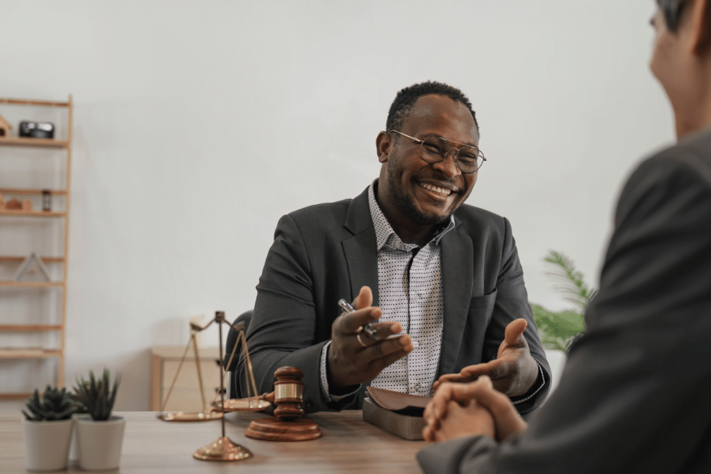 A smiling man in a suit sits at a desk discussing the industries we serve with another person. He holds a pen, and a small notebook is in front of him. Legal symbols, including a gavel and scales, adorn the desk. A shelf and plants are arranged in the background.