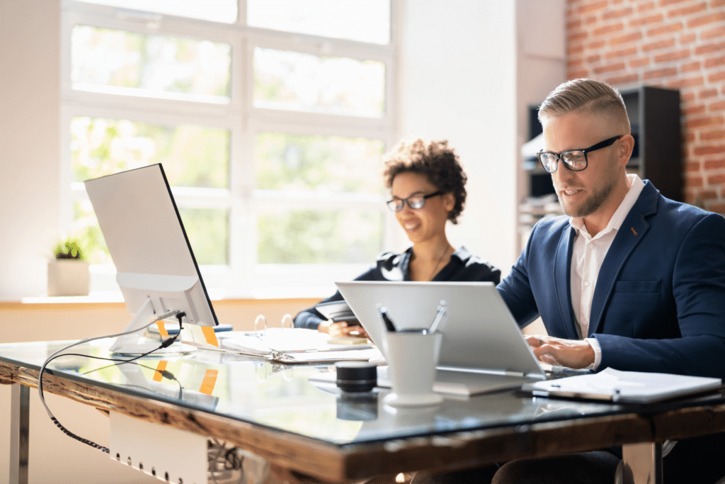 Two people work at a desk in a bright office, immersed in tasks from various industries. One uses a laptop, the other a desktop computer. Both appear focused and are wearing glasses. The glass-top desk displays several office items, while large windows grace the background.