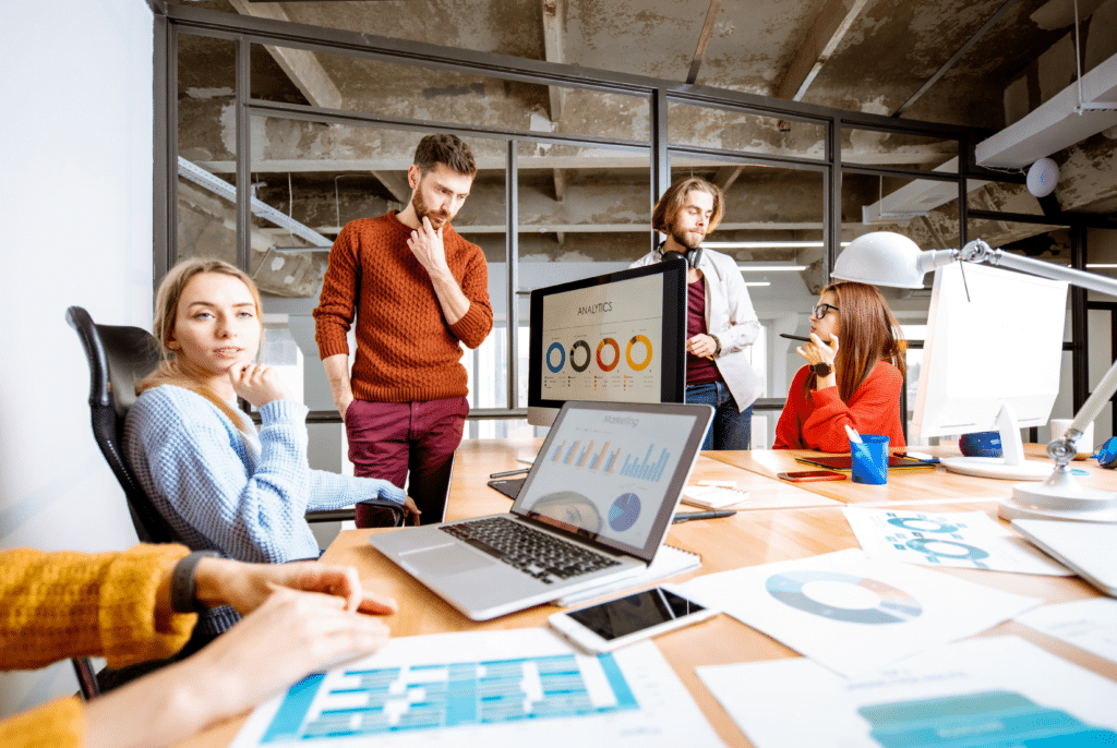 A group of people in a modern office setting is gathered around a table with multiple laptops and documents displaying industry-specific charts. They appear engaged in a discussion or presentation, while a large screen showcases a graph titled "Industries We Serve: Analytics.