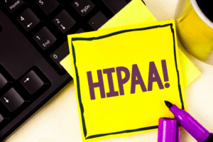 A sticky note with HIPAA! written in bold purple letters sits on a laptop keyboard, symbolizing the importance of HIPAA-compliant IT services. Two purple markers lie nearby, while the edge of a yellow mug peeks from the corner, emphasizing data security and compliance.