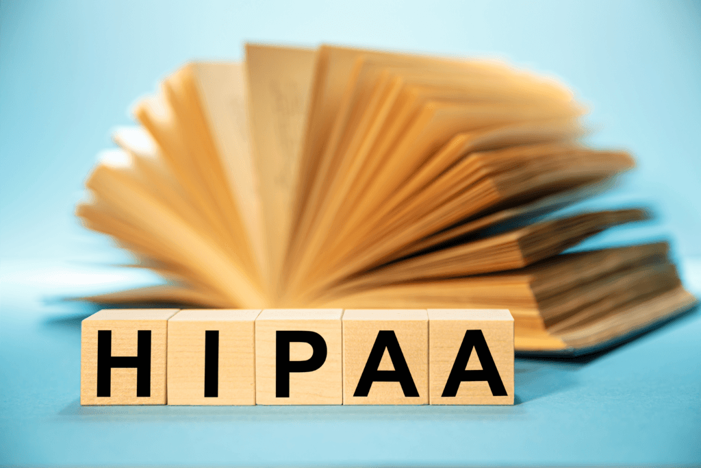 Wooden blocks spelling HIPAA are in the foreground, emphasizing the importance of HIPAA-compliant IT services. Behind them, an open book with blurred pages is set against a blue background, symbolizing the vast knowledge needed for ensuring data security and compliance.