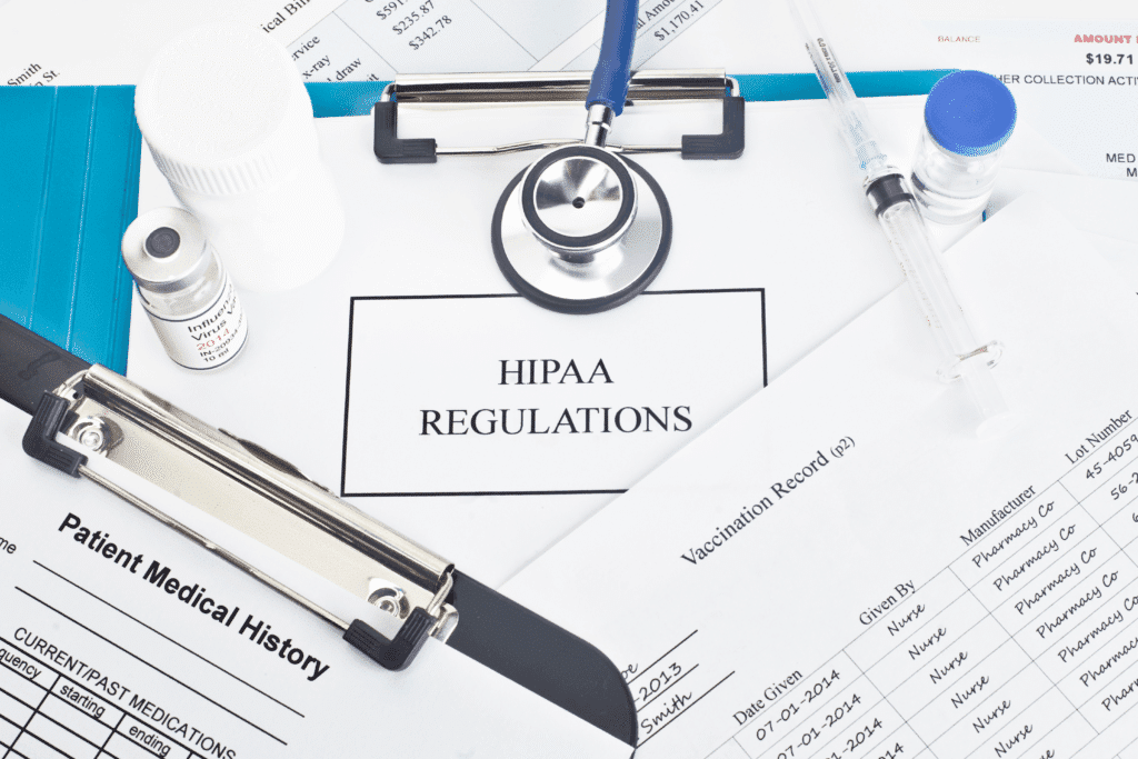 A medical setup features a stethoscope, vials, syringe, and documents labeled HIPAA Regulations, Patient Medical History, and Vaccination Record on a clipboard. Nearby are medical forms and a pen, all safeguarded by HIPAA-compliant IT services to ensure data security and compliance.