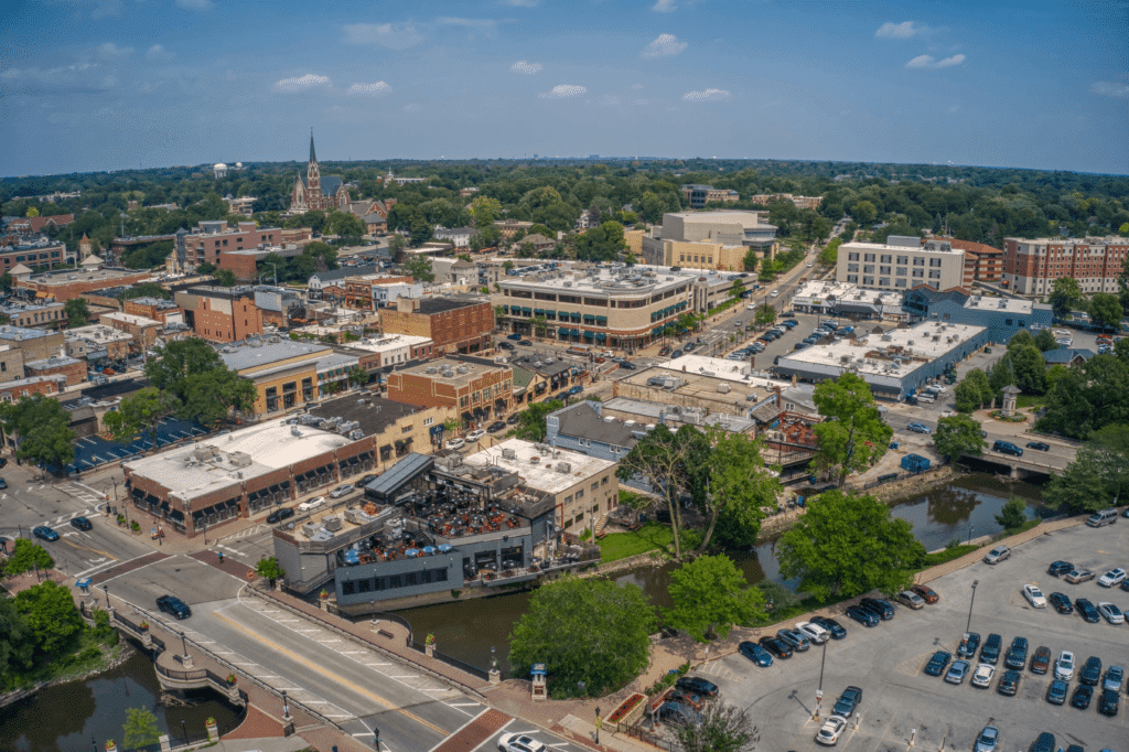 Aerial view of a small town featuring a mix of commercial buildings and IT services in Naperville, with streets and a church steeple. Trees and a river wind through the area, cars parked near the bridge and along roads. Bright, clear weather with scattered clouds.