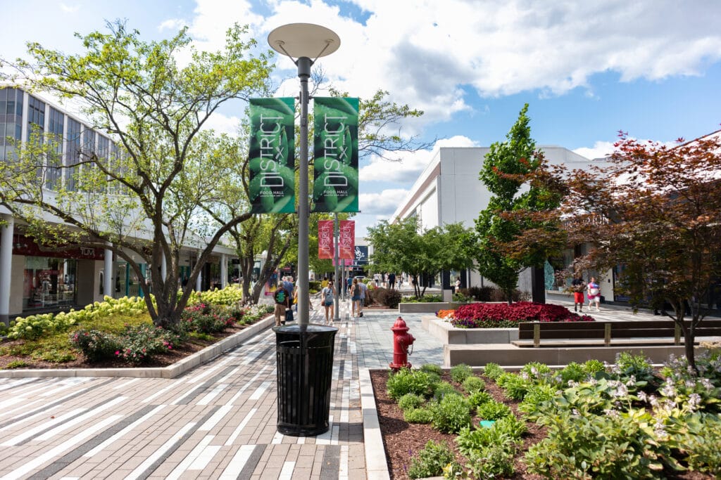 A vibrant outdoor shopping district in Glenview boasts greenery and modern architecture. People stroll the brick-paved walkway flanked by colorful flower beds and trees, where hints of IT services banners hang from lampposts under a partly cloudy sky.