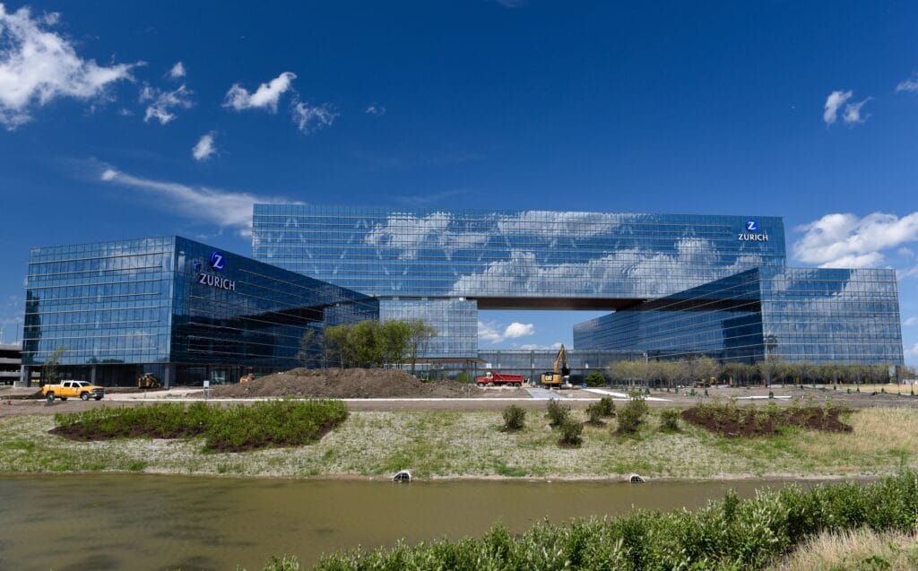 A large, modern glass building with the word Zurich on it reflects a blue sky with clouds. This unique structure, reminiscent of innovative designs associated with IT services in Schaumburg, features a central open space. In the foreground is a landscaped area and a construction site.