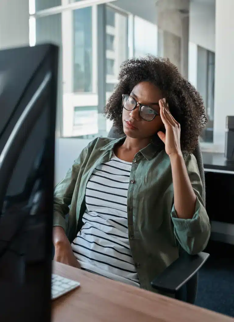 A woman wearing glasses and a striped shirt sits at a desk in an office, looking tired or stressed as she rests her head on her hand and gazes at a computer monitor—highlighting the need for reliable Business IT Support.